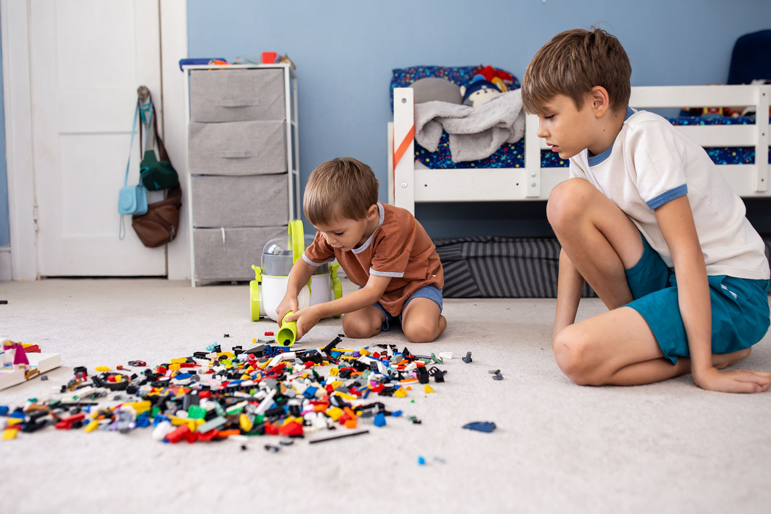 A young boy uses his Pick-Up Bricks toy vacuum to clean up a mess of LEGO spread across the floor as his older brother watches